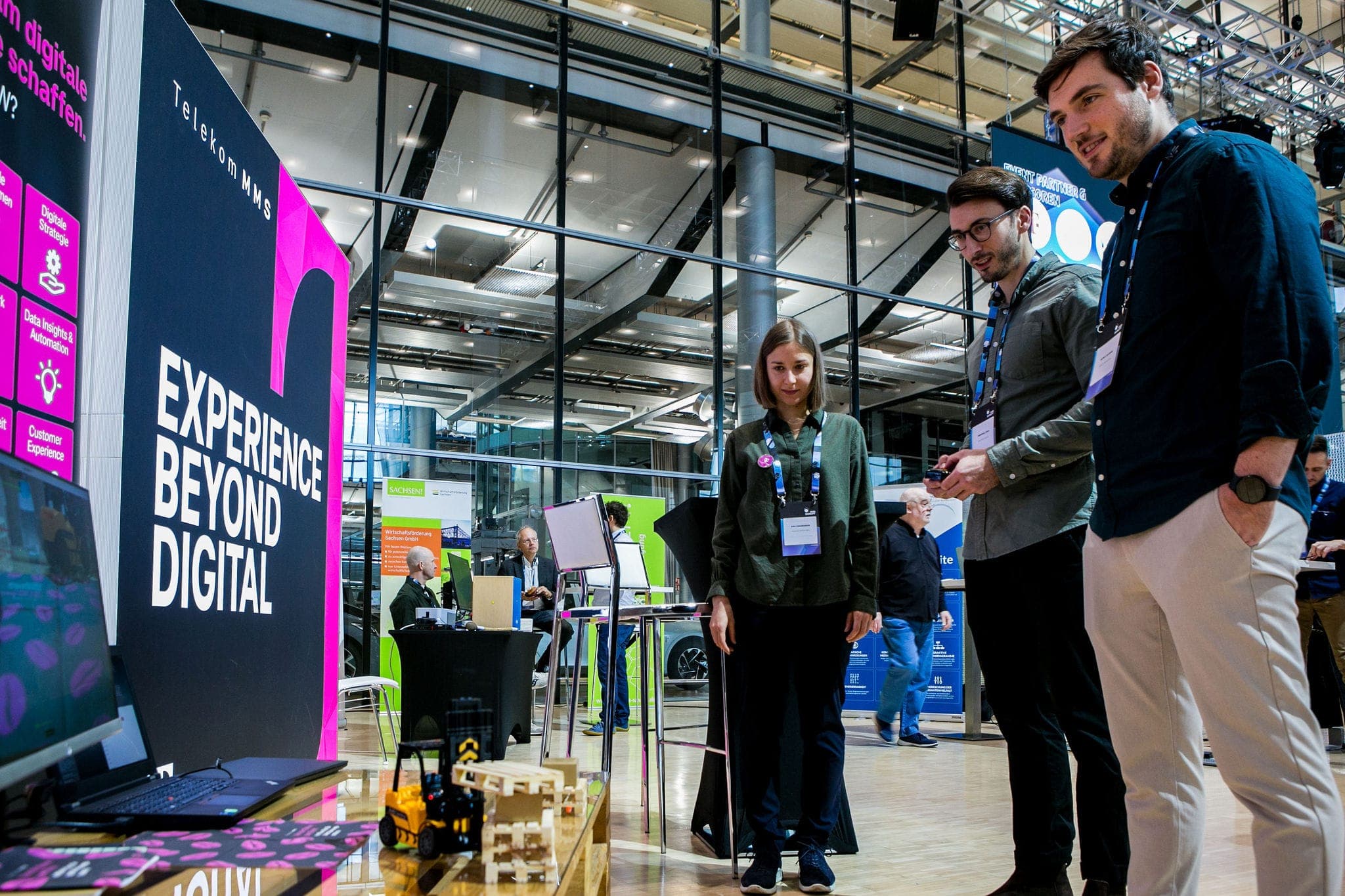 Ausstellerstand der Telekom MMS mit drei Personen, eine Person steuert mit einen Controller einen Mini-Gabelstapler.